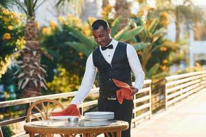 Wipes plates by red cloth. Black waiter in formal clothes is at his work outdoors at sunny daytime photo