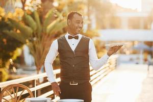 Holds empty plate. Black waiter in formal clothes is at his work outdoors at sunny daytime photo