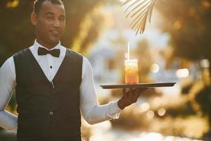 With order in hand. Black waiter in formal clothes is at his work outdoors at sunny daytime photo