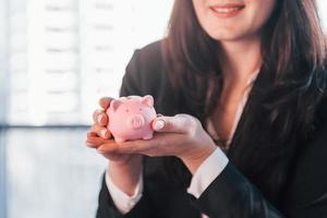 Woman in black formal clothes is indoors in the modern office holds pink piggy bank photo