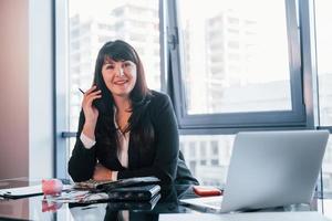 con laptop en la mesa. mujer con ropa formal negra está adentro en la oficina moderna foto