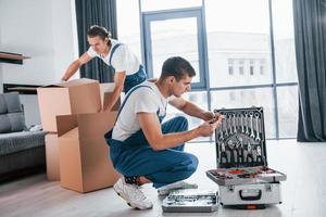 Uses tools from case. Two young movers in blue uniform working indoors in the room photo