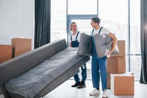 Carrying heavy sofa. Two young movers in blue uniform working indoors in the room photo