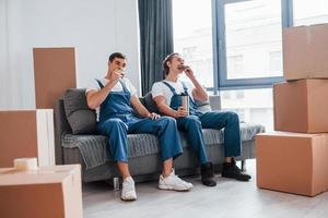 Sitting on the sofa. Tired and taking break. Two young movers in blue uniform working indoors in the room photo