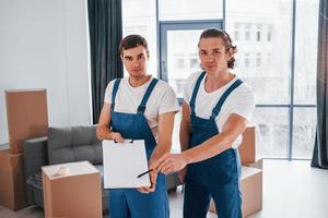 Holds notepad with document. Two young movers in blue uniform working indoors in the room photo