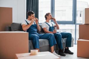 Sitting on the sofa. Tired and taking break. Two young movers in blue uniform working indoors in the room photo