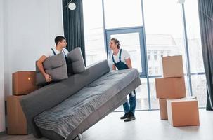 Carrying heavy sofa. Two young movers in blue uniform working indoors in the room photo