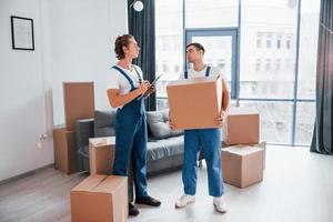 Holds notepad with document. Two young movers in blue uniform working indoors in the room photo