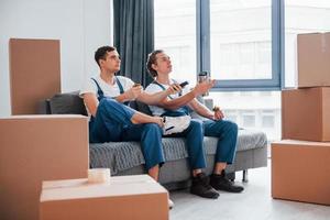 Sitting on the sofa. Tired and taking break. Two young movers in blue uniform working indoors in the room photo