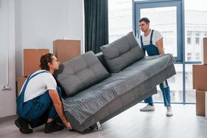 llevar un sofá pesado. dos jóvenes motores con uniforme azul trabajando en el interior de la habitación foto