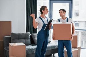 Holds notepad with document. Two young movers in blue uniform working indoors in the room photo