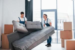 Carrying heavy sofa. Two young movers in blue uniform working indoors in the room photo