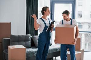 Holds notepad with document. Two young movers in blue uniform working indoors in the room photo