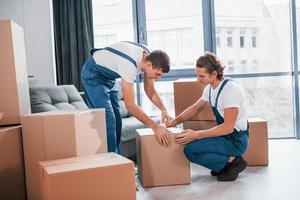 Packaging the box. Two young movers in blue uniform working indoors in the room