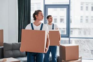 With boxes in hands. Two young movers in blue uniform working indoors in the room photo