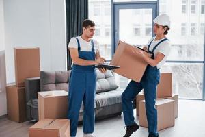 Holds notepad with document. Two young movers in blue uniform working indoors in the room photo