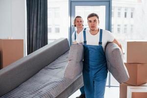 Carrying heavy sofa. Two young movers in blue uniform working indoors in the room photo