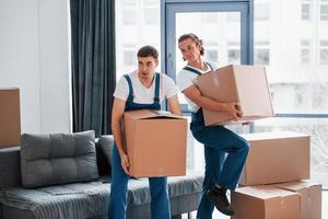 With boxes in hands. Two young movers in blue uniform working indoors in the room photo