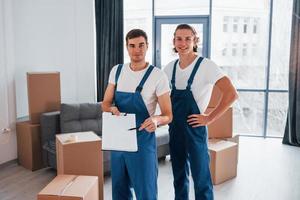 Holds notepad with document. Two young movers in blue uniform working indoors in the room photo