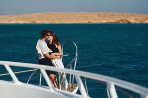 Kissing each other. Cheerful young couple standing on the yacht on the sea. Conception of vacation photo
