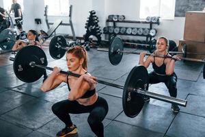 Squats with barbell. Group of young sportive people have crossfit day indoors in gym photo