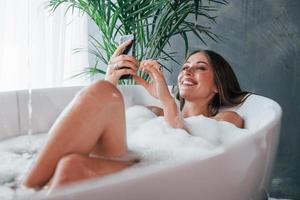 Uses phone. Beautiful young woman have free time at home in the bath photo