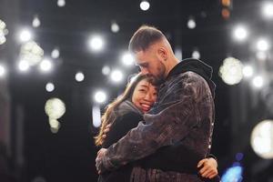 abrazándose unos a otros. feliz pareja multirracial juntos al aire libre en la ciudad celebrando el año nuevo foto