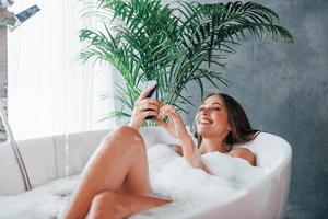 usa teléfono. bella joven tiene tiempo libre en casa en el baño foto