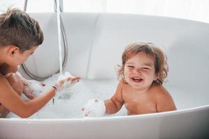 Two kids having fun and washing themselves in the bath at home. Posing for a camera photo