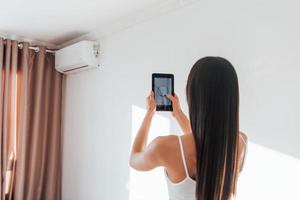 vista trasera de la mujer que está de pie en el interior con el teléfono y controla la tecnología de la casa inteligente foto