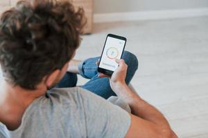 Sits and holds phone in hands. Rear view of adult man that is indoors controlling smart home technology photo