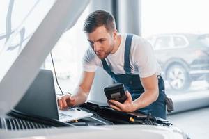 Uses laptop. Young man in white shirt and blue uniform repairs automobile photo