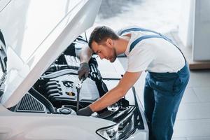 Hood is opened. Young man in white shirt and blue uniform repairs automobile photo