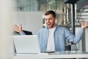 Uses laptop. Young man in white shirt and blue jacket works indoors photo