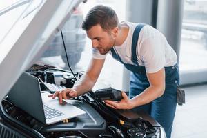 utiliza portátil. joven con camisa blanca y uniforme azul repara automóvil foto