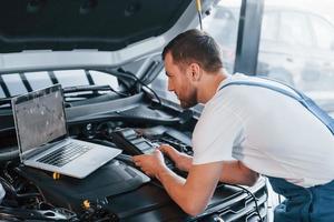 Uses laptop. Young man in white shirt and blue uniform repairs automobile photo
