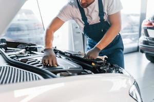 Quality service. Young man in white shirt and blue uniform repairs automobile photo