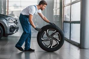 Replacement of wheel. Young man in white shirt and blue uniform repairs automobile photo