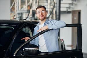 Successful man in glasses standing near brand new car indoors photo