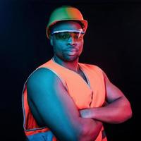 Construction worker in uniform and hard hat. Futuristic neon lighting. Young african american man in the studio photo