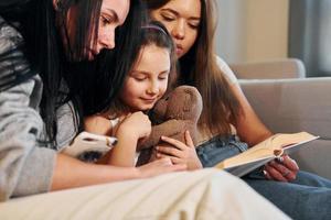 Reading book together. Female lesbian couple with little daughter spending time together at home photo