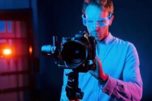 Holds camera. Neon lighting. Young european man is in the dark studio photo