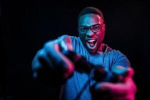 Plays game by using controller. Futuristic neon lighting. Young african american man in the studio photo