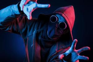 Scary guy in eyewear, mask and hood. Neon lighting. Young european man is in the dark studio photo