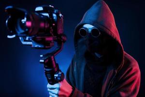 Guy in hood with camera. Neon lighting. Young european man is in the dark studio photo