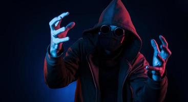 Scary guy in eyewear, mask and hood. Neon lighting. Young european man is in the dark studio photo