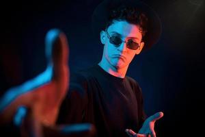 In hat and glasses. Neon lighting. Young european man is in the dark studio photo