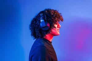 With headphones. Young beautiful man with curly hair is indoors in the studio with neon lighting photo