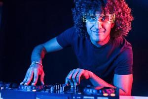 hombre con cabello rizado usando equipo de dj y parado en la oscura habitación iluminada con neón foto