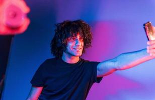 Takes selfie. Young beautiful man with curly hair is indoors in the studio with neon lighting photo